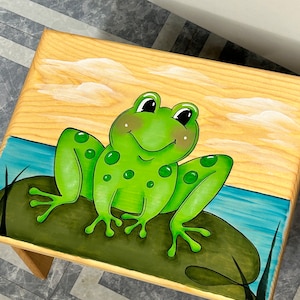 May include: A wooden step stool featuring a hand-painted frog design. The frog is green with black eyes and sits on a green lily pad. The background depicts a blue water scene with a yellow sky and white clouds. The stool has four wooden legs.