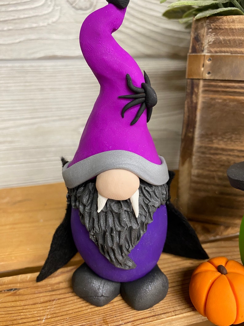 May include: Purple and black Halloween gnome with a pointy hat, a spider on the hat, a black cape, and fangs.