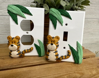 May include: Two white light switch covers featuring a tiger and jungle leaf design. The left cover has a double outlet and the right cover has a single switch.