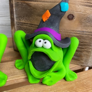 May include: A green frog wearing a black witch hat with a purple band and orange and blue accents. The frog has a wide open mouth and large, round eyes.