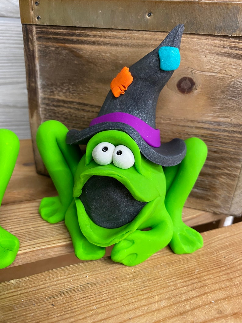 May include: A green frog wearing a black witch hat with a purple band and orange and blue accents. The frog has a wide open mouth and large, round eyes.