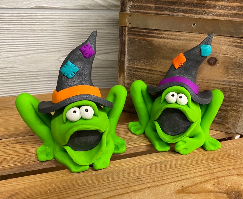 May include: Two green frog figurines wearing black witch hats with orange and purple accents. The frogs have large, round eyes and open mouths.