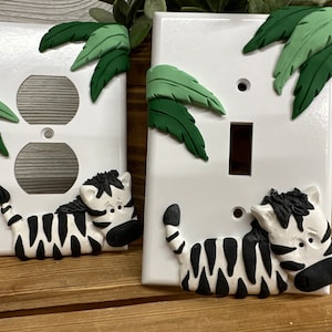 May include: Two white light switch covers with a zebra and green leaves design. The zebra is black and white and is laying down. The leaves are green and are on the top of the switch covers.