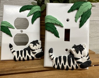 May include: Two white light switch covers with a zebra and green leaves design. The zebra is black and white and is laying down. The leaves are green and are on the top of the switch covers.