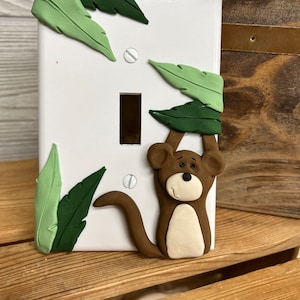 May include: A white light switch cover with a brown monkey hanging from green leaves. The monkey has a white face and a brown body.