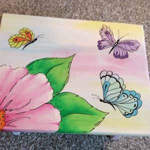 May include: Hand-painted wooden box featuring a pink flower, green leaves, and three butterflies. The butterflies are yellow, purple, and blue.
