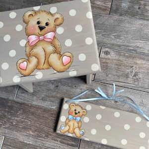 May include: A wooden stool with a light brown base and a light gray top. The top has a white polka dot pattern and a painted image of a brown teddy bear with a pink bow. The stool has a matching sign with a similar design.