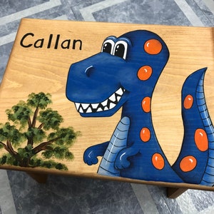 May include: Wooden step stool with a hand-painted blue dinosaur design and the name "Callan" in black. The dinosaur has orange spots and a friendly expression. A green tree is painted on the side.