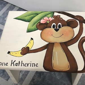 May include: A white wooden stool with a hand-painted design of a brown monkey holding a yellow banana. The monkey has a pink flower in its hair and is smiling. The stool is personalized with the name "Anne Katherine".