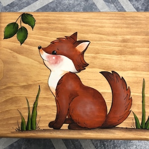 May include: A wooden box with a painted image of a fox looking up at a green leaf branch. The fox is brown with a white chest and a bushy tail. The background is a light brown wood.