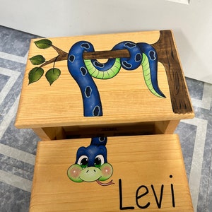 May include: A wooden two-step stool painted with a blue snake wrapped around a tree branch. The bottom step has a cartoon snake face with a green tongue and the name "Levi" written in black.