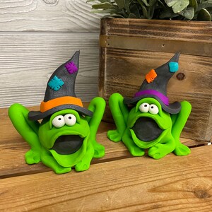 May include: Two green frog figurines wearing black witch hats with colorful accents. The frogs have large eyes and open mouths.