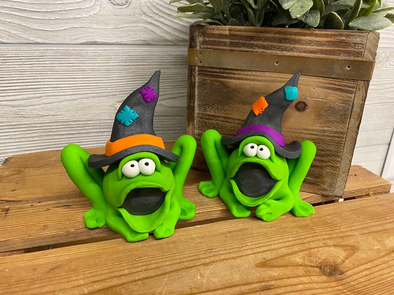 May include: Two green frog figurines wearing black witch hats with colorful accents. The frogs have large eyes and open mouths.