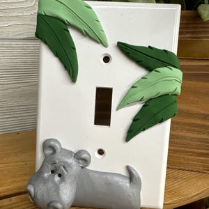 May include: A white light switch cover with a gray hippopotamus and green leaves sculpted on it.
