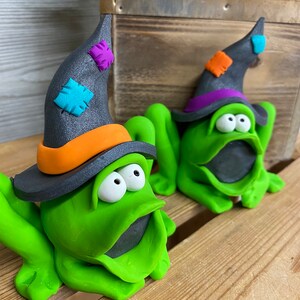 May include: Two green frog figurines wearing black witch hats with colorful patches. The frogs have large eyes and open mouths. The hats have orange and purple trim.