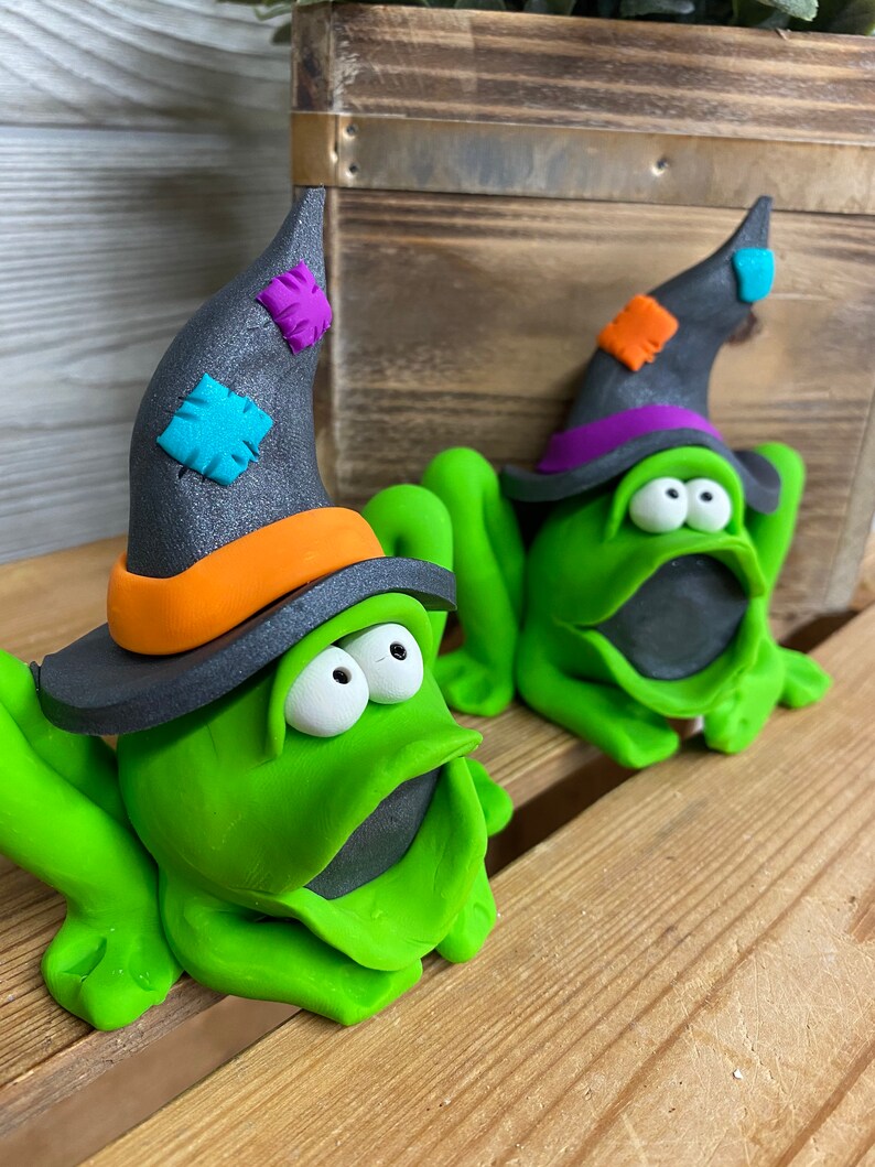 May include: Two green frog figurines wearing black witch hats with colorful patches. The frogs have large eyes and open mouths. The hats have orange and purple trim.