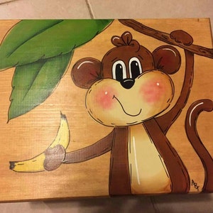 May include: A wooden box with a hand-painted design of a monkey holding a banana. The monkey is brown with a light brown face and a white stripe down its back. The banana is yellow with brown spots. The box is painted with a green leaf design.