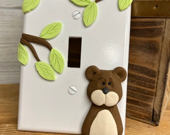 May include: A white light switch cover with a brown bear and green leaves sculpted on it.
