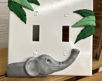 May include: A white light switch cover featuring a grey elephant and green leaves. The elephant's trunk is raised and the leaves are positioned around the switch.