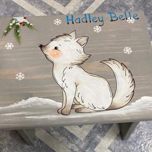 May include: A gray wooden stool with a white arctic fox painted on it. The fox is sitting on a snowy background with snowflakes and a sprig of pine with red berries. The stool is personalized with the name "Hadley Belle" in blue lettering.