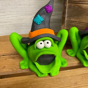 May include: A green frog wearing a black witch hat with orange and purple accents. The frog has large white eyes and a wide open mouth.