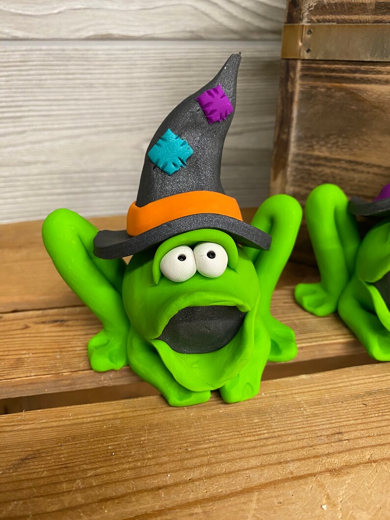 May include: A green frog wearing a black witch hat with orange and purple accents. The frog has large white eyes and a wide open mouth.