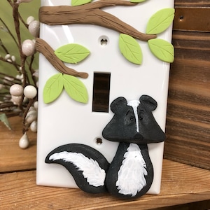 May include: A white light switch cover with a 3D sculpted design of a black and white skunk with a brown tree branch and green leaves.