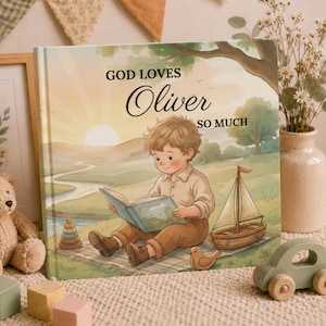 Op de afbeelding: Een vierkant hardcover boek met de titel GOD LOVES Oliver SO MUCH, met een illustratie van een jong kind dat op een kleed buiten een boek leest, met een speelgoedzeilboot en een stapelspeeltje in een landschap met zachte pastelkleuren.