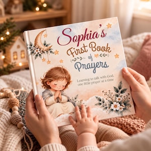 May include: A hardcover book titled Sophia's First Book of Prayers, Learning to talk with God, one little prayer at a time. The cover features a watercolor illustration of a child praying, a crescent moon, stars, and white flowers.