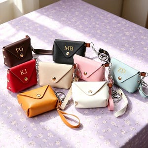 May include: A collection of small, envelope-style pouches in various colors including brown, black, red, cream, pink, yellow, white, and light blue. Each pouch features a gold-tone snap closure, a wrist strap, and gold-tone monogrammed initials.