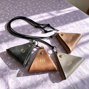 May include: Four triangular coin pouches in black, tan, light brown, and grey, each featuring a gold-tone snap button and gold-stamped initials KJ, FG, ZD, and MB. Each pouch is attached to a long black cord with a metal ring for carrying.