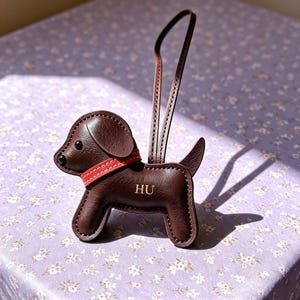 Puede incluir: Un llavero de cuero marrón oscuro con forma de perro, un collar rojo brillante, ojos de cuentas negras y las letras HU grabadas en oro en el costado. El accesorio incluye una correa de cuero larga y fina y costuras blancas visibles.