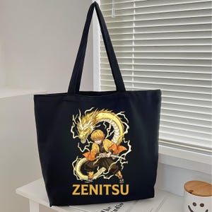 May include: A black tote bag featuring a graphic print of a blonde character in a yellow and orange patterned haori holding a sword, surrounded by a yellow lightning dragon. The name ZENITSU is printed in bold yellow capital letters below the graphic.