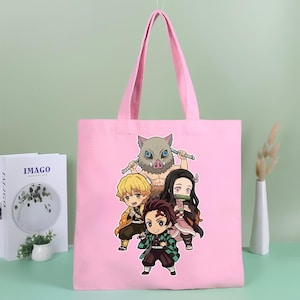 May include: A light pink tote bag featuring a colorful graphic print of four anime characters in a chibi style. The design includes a boar-headed figure, a blonde character, a dark-haired character with a bamboo muzzle, and a central character.