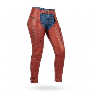May include: Red leather chaps worn over blue denim jeans. The chaps feature a buckled waist belt, vertical zippered pockets on the upper thighs, and front-facing lace-up detailing along the length of each leg, ending with adjustable ankle straps.