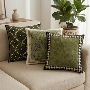 Puede incluir: Tres cojines decorativos sobre un sofá beige. Los cojines presentan varios diseños, incluyendo florales y geométricos, en tonos verdes, negros y blancos. Uno de los cojines tiene el texto "Romantic Garden" impreso.