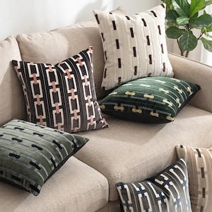 Può includere: Una collezione di cuscini quadrati caratterizzati da un motivo geometrico ripetuto a maglie di catena. I cuscini sono realizzati in diverse combinazioni di colori, tra cui crema con marrone, verde scuro con giallo e grigio con accenti rosa o blu.