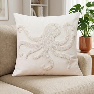Peut inclure: Un coussin carré présentant un motif de pieuvre blanc, texturé et touffeté, centré sur une housse en tissu blanc cassé lisse. Le motif de pieuvre est réalisé avec du fil bouclé en relief, offrant un aspect tactile et tridimensionnel sur le fond plat du coussin.