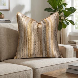 Può includere: Cuscino quadrato decorativo con un motivo a righe verticali dorate, beige e grigie. Il cuscino ha un bordo dorato ed è posto su un divano beige. Questo cuscino decorativo aggiunge un tocco di eleganza al soggiorno.