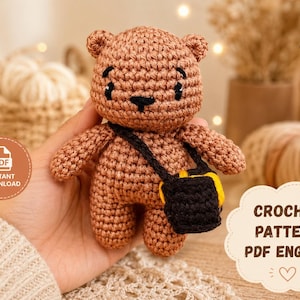May include: A small brown crocheted bear toy featuring a black crossbody bag with yellow accents. The bear has embroidered black eyes and a black nose. A circular badge reads PDF INSTANT DOWNLOAD and a cloud-shaped graphic says CROCHET PATTERN PDF ENGLISH.