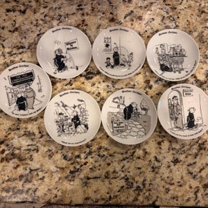 May include: A set of seven white ceramic plates, each featuring a black and white cartoon illustration of Brother Juniper with unique captions. The plates display various humorous scenes, including a tailor shop, a grocery store, and garden settings.