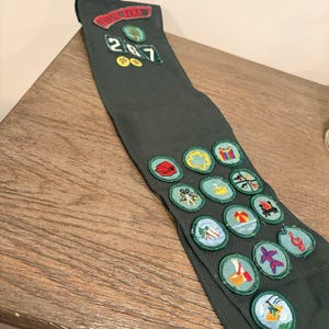 May include: A dark green sash adorned with numerous embroidered circular merit badges featuring various colorful designs. At the top, a red rectangular patch reads "BASIN BEND" above the numbers "2", "6", and "7" in white on green.