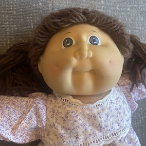 May include: A doll with a soft, rounded face, blue eyes, and brown yarn hair styled in pigtails. The doll wears a white dress featuring a small purple floral pattern and delicate white lace trim along the neckline and bodice area.