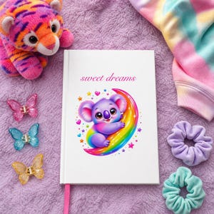 A white journal with a rainbow koala on a crescent moon, and the text "sweet dreams", on a purple fuzzy blanket with a tiger plush, butterfly clips, and scrunchies.