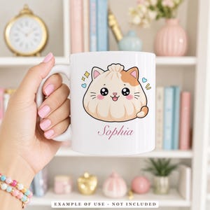 A hand holding a white mug with a cartoon illustration of a smiling dumpling with cat ears and the name "Sophia" written below it.