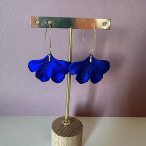 Op de afbeelding: Een paar helderblauwe oorbellen in de vorm van ginkgoblaadjes. De oorbellen worden gepresenteerd op een goudkleurige standaard met een houten voet. De achtergrond is zachtroze.