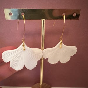 Peut inclure: Une paire de boucles d'oreilles blanches en forme de pétale avec des crochets et des accents dorés. Les boucles d'oreilles sont présentées sur un présentoir à boucles d'oreilles doré sur fond marron. Surface texturée.