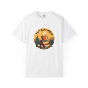 Peut inclure: T-shirt blanc avec un motif circulaire portant l'inscription "COZY GAME NIGHT". Le motif comprend une tasse de café, des livres, des dés et une scène de forêt. Le t-shirt est en matière souple.