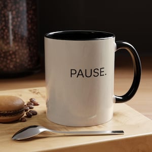 May include: A cream-colored coffee mug with a black interior and handle. The word "PAUSE." is printed in black on the front. A silver spoon, a macaron, and coffee beans are on a wooden surface.