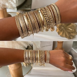 May include: A stack of multiple gold-toned bracelets worn on the wrist, featuring a variety of textures including smooth polished bands, twisted rope designs, chain links, and thin bangles with small dangling charms attached to several pieces.